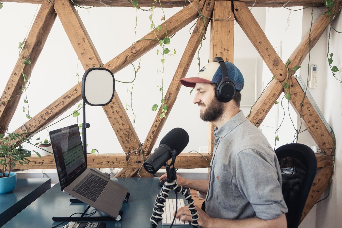A presenter at a laptop and microphone in a bright studio with timber beams and plants