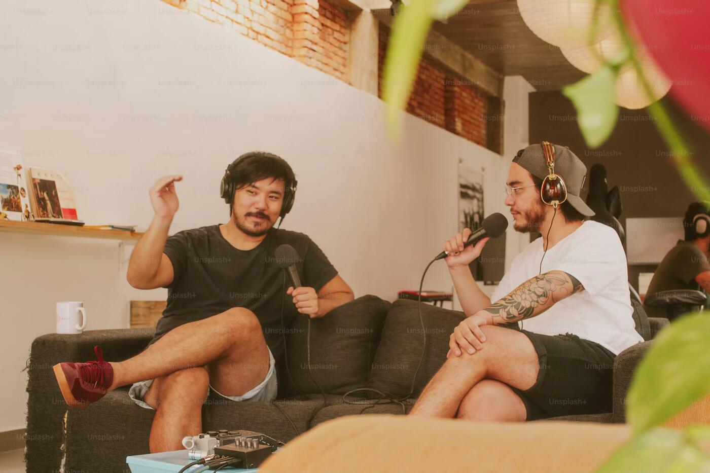 Two podcasters laughing and recording in a relaxed studio with brick walls and plants